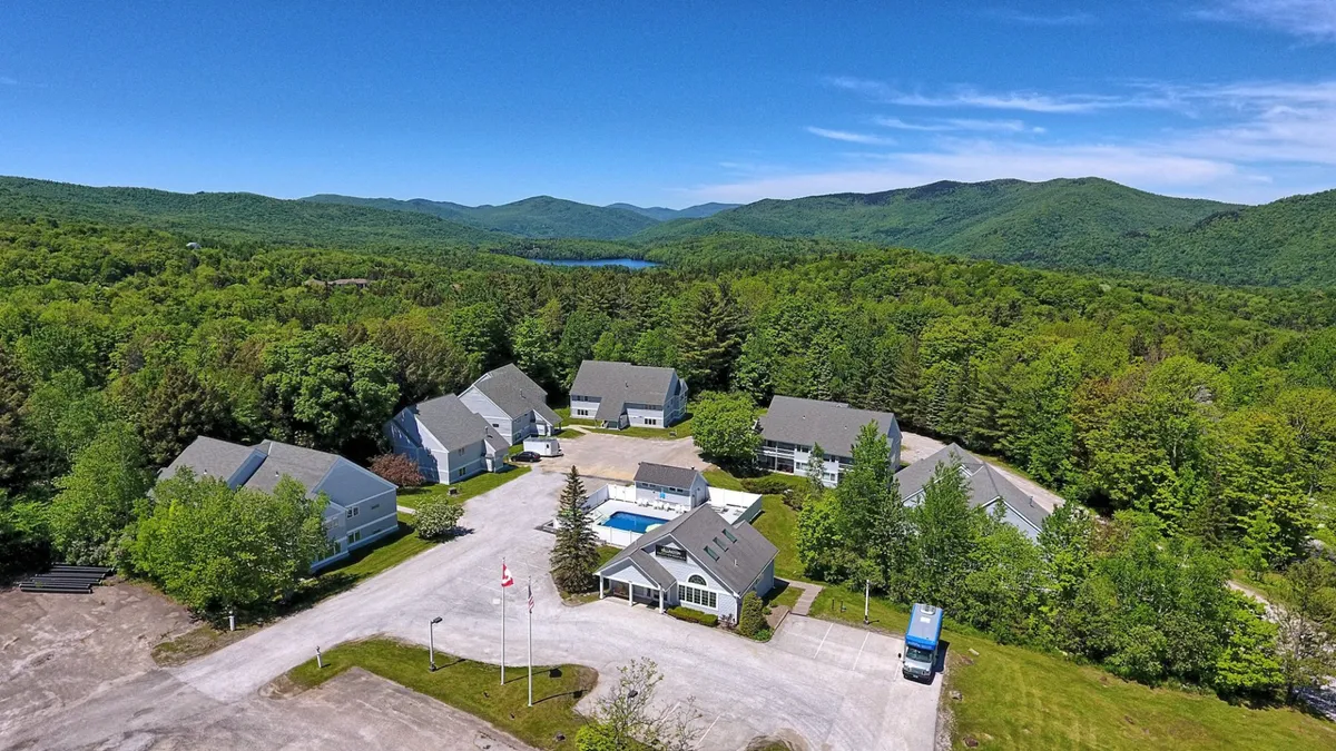 This exterior image features the main building set against a backdrop of mountains and a lake, with a swimming pool and outdoor areas surrounded by lush trees. The serene setting hints at proximity to natural attractions and the outdoor adventure opportunities that abound in the region.