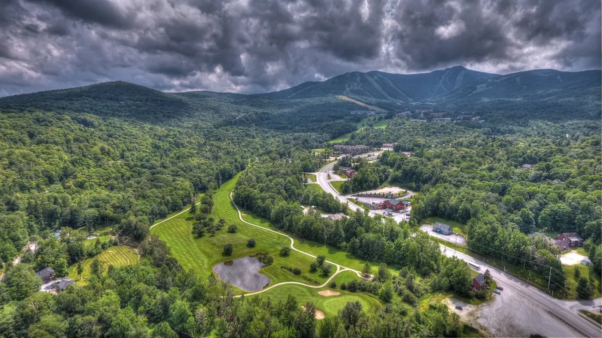 A panoramic view of tree-covered mountains, a tranquil pond, a sprawling golf course, and dense Vermont forest. The landscape promises outdoor adventure and natural beauty, signaling the area’s recreational offerings.