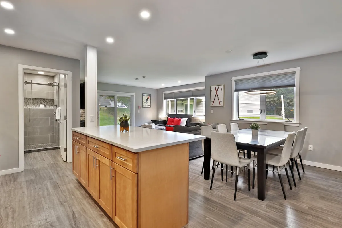 A broad angle of the open living area, showing the kitchen island with bar seating, adjacent dining table, and a cozy living area with a sofa. The setup is ideal for cooking, dining, and relaxing together, with elements like the kitchen island and dining table both visible for a sense of flow and versatility.