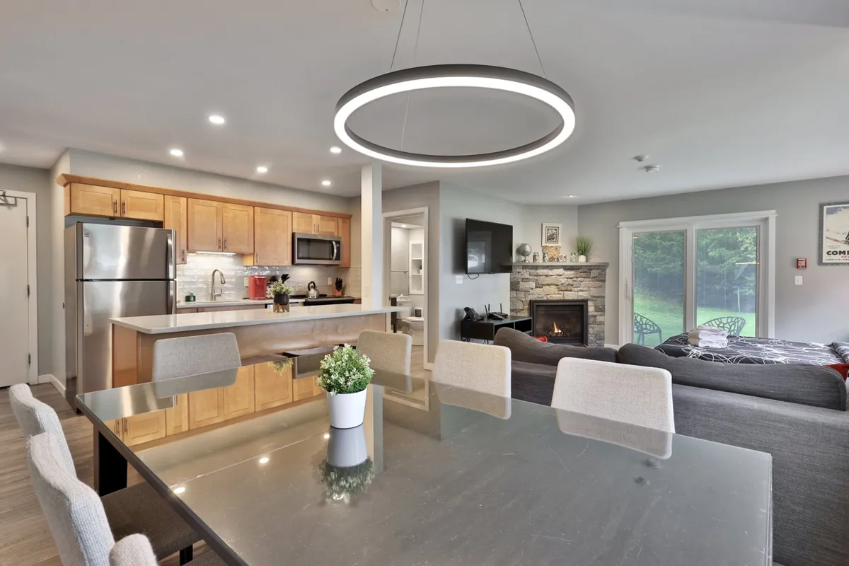 The image showcases a stylish open-concept living and dining area with a modern kitchen island, an inviting dining table ideal for group meals or gatherings, a cozy fireplace, and large windows that flood the space with natural light. The contemporary light fixture adds warmth and elegance, creating an atmosphere perfect for relaxing after a day on the slopes or enjoying time with family and friends.