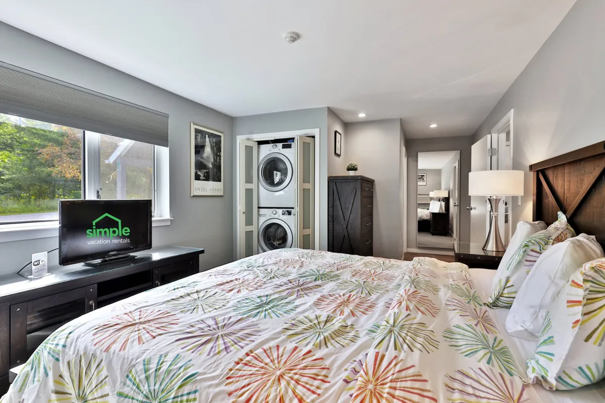 A cozy bedroom featuring a king-sized bed with colorful bedding, a smart TV on the dresser, and a closet with in-unit washer and dryer visible. The scene exudes warmth, comfort, and convenience, ideal for extended stays or restful nights.