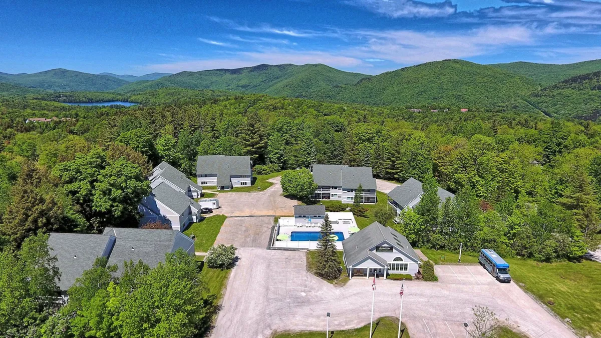A wide exterior shot of the inn’s main lodge and the expansive seasonal swimming pool, surrounded by lounge chairs, umbrellas, and views of the rolling mountains in the background. The scene feels calm and inviting, with hints of lush landscaping and ample space for guests to relax or socialize.
