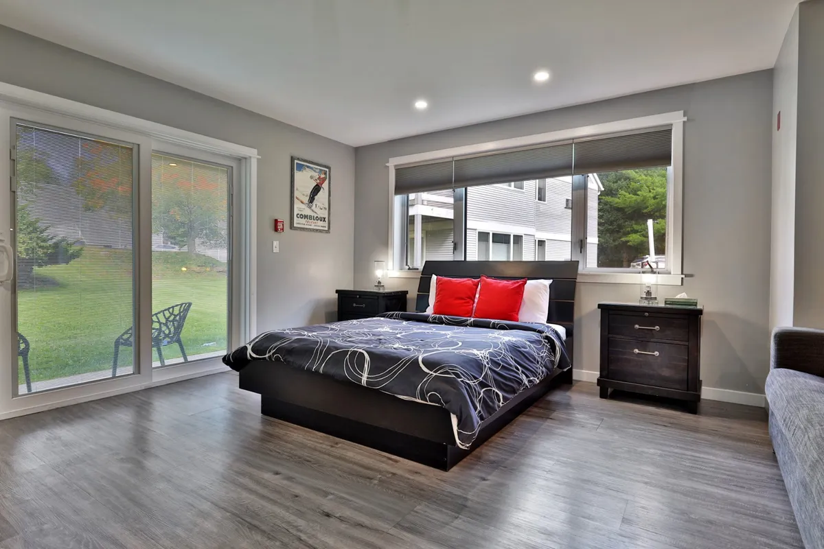 This image shows a serene bedroom with a bed dressed in patterned bedding, red accent pillows, and large windows that let in natural light. Nightstands flank the bed, adding to the cozy and inviting atmosphere. The setup suggests a restful space with beautiful morning views, ideal for couples.