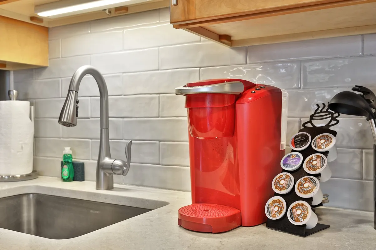 A close-up view of a kitchen counter featuring a red coffee maker, sink, and kitchen utensils, with a coffee pod holder nearby. The setting is practical and inviting, suggesting both convenience and hospitality for guests.