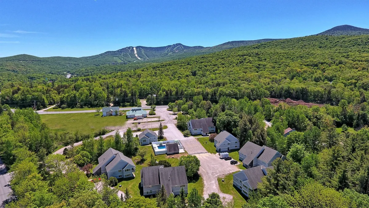 An exterior scene featuring the swimming pool nestled among greenery, mountain views in the distance, and adjacent cabins or accommodations. The relaxed setting includes people enjoying swimming, lounging, or nature activities, emphasizing the community and outdoor lifestyle offered.